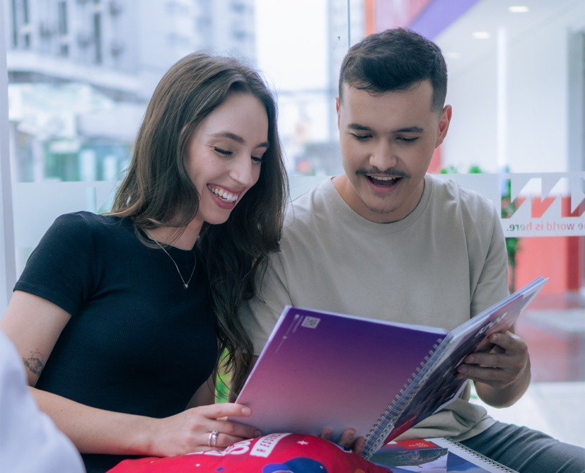 Home Dois jovens estudantes sorridentes consultando material didático na recepção da KNN Idiomas Pinhais.
